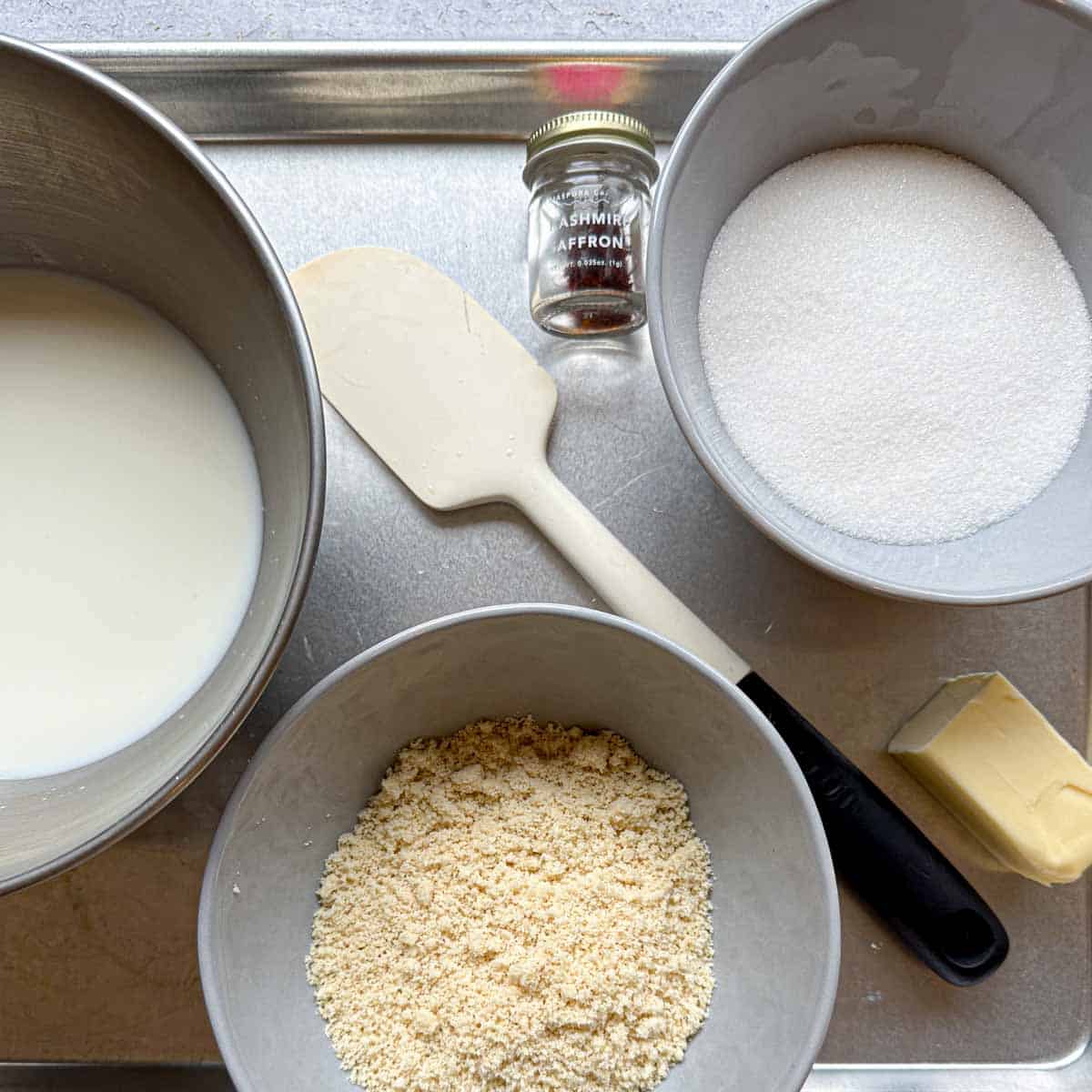 Ingredients for badam halwa on a sheet tray. Milk, almond flour, butter, sugar, saffron, and rubber spatula.