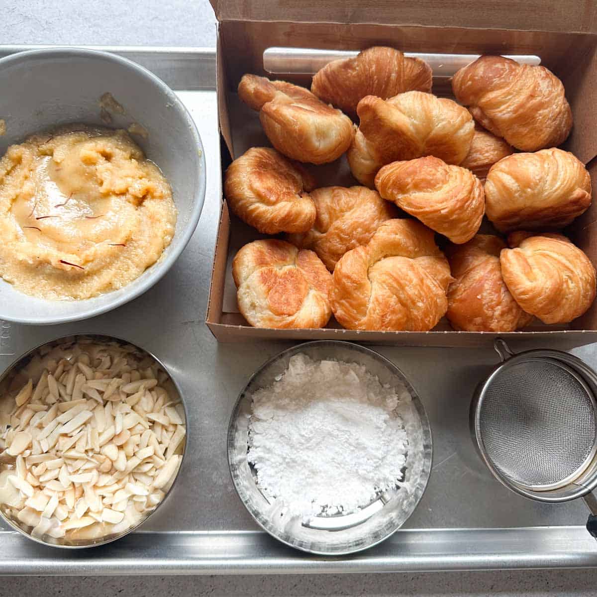 Ingredients for badam halwa croissants on a sheet tray. It includes slivered almonds, badam halwa, powdered sugar, and mini croissants.