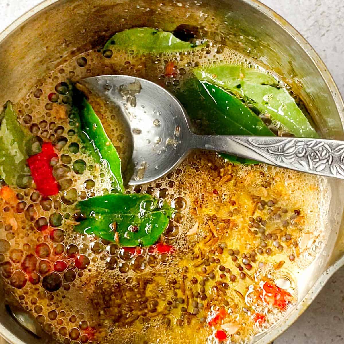 Ghee sizzling in a saucepan with cumin seeds, mustard seeds, fresh chili, and curry leaves