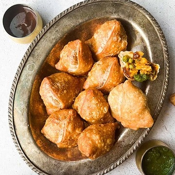 Samosas on a platter with tamarind chutney on top left and cilantro mint chutney on bottom right. One samosa is broken open revealing the potato and peas filling.