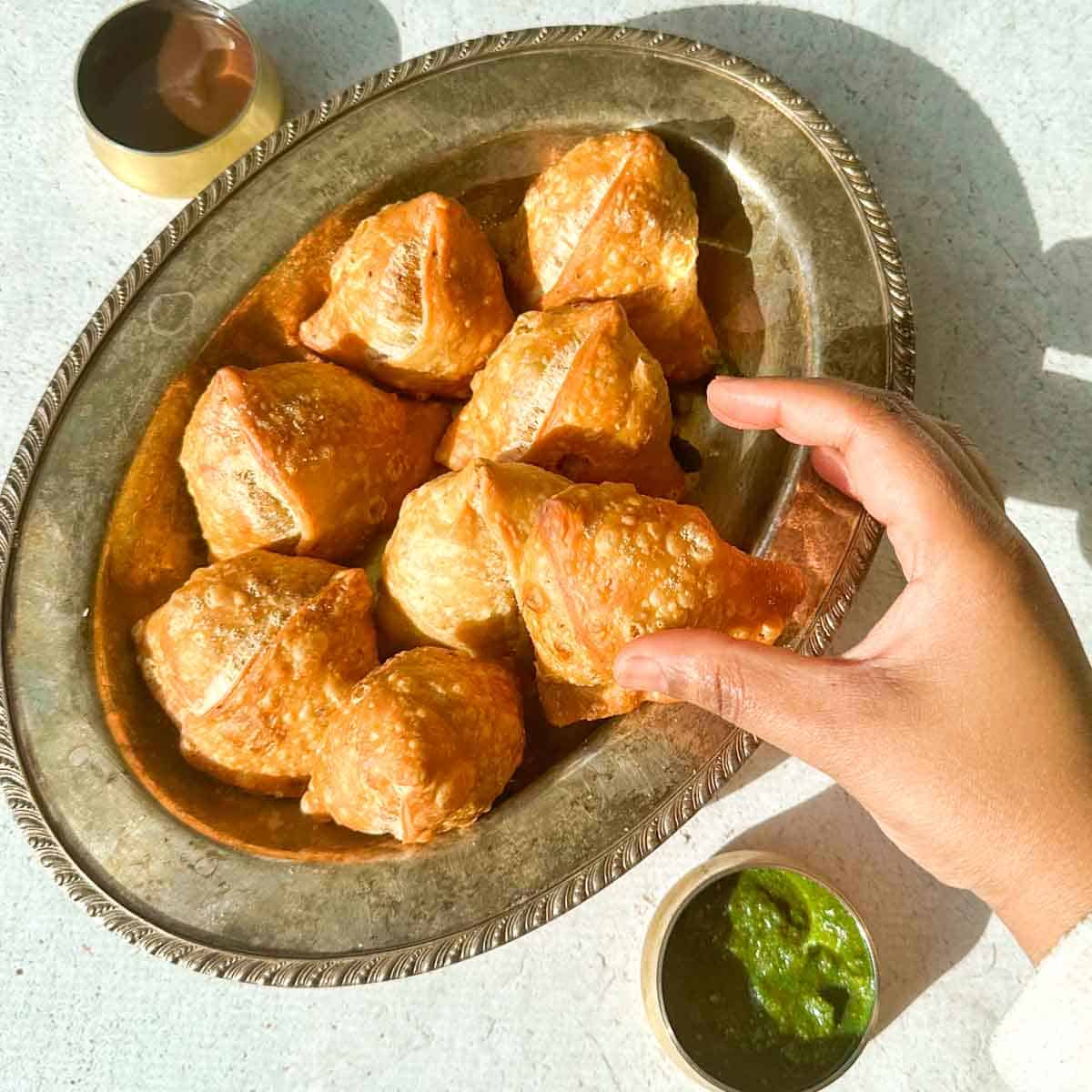A hand grabbing a samosa from a platter of samosas. Green chutney on the bottom right and tamarind date chutney on the top left.