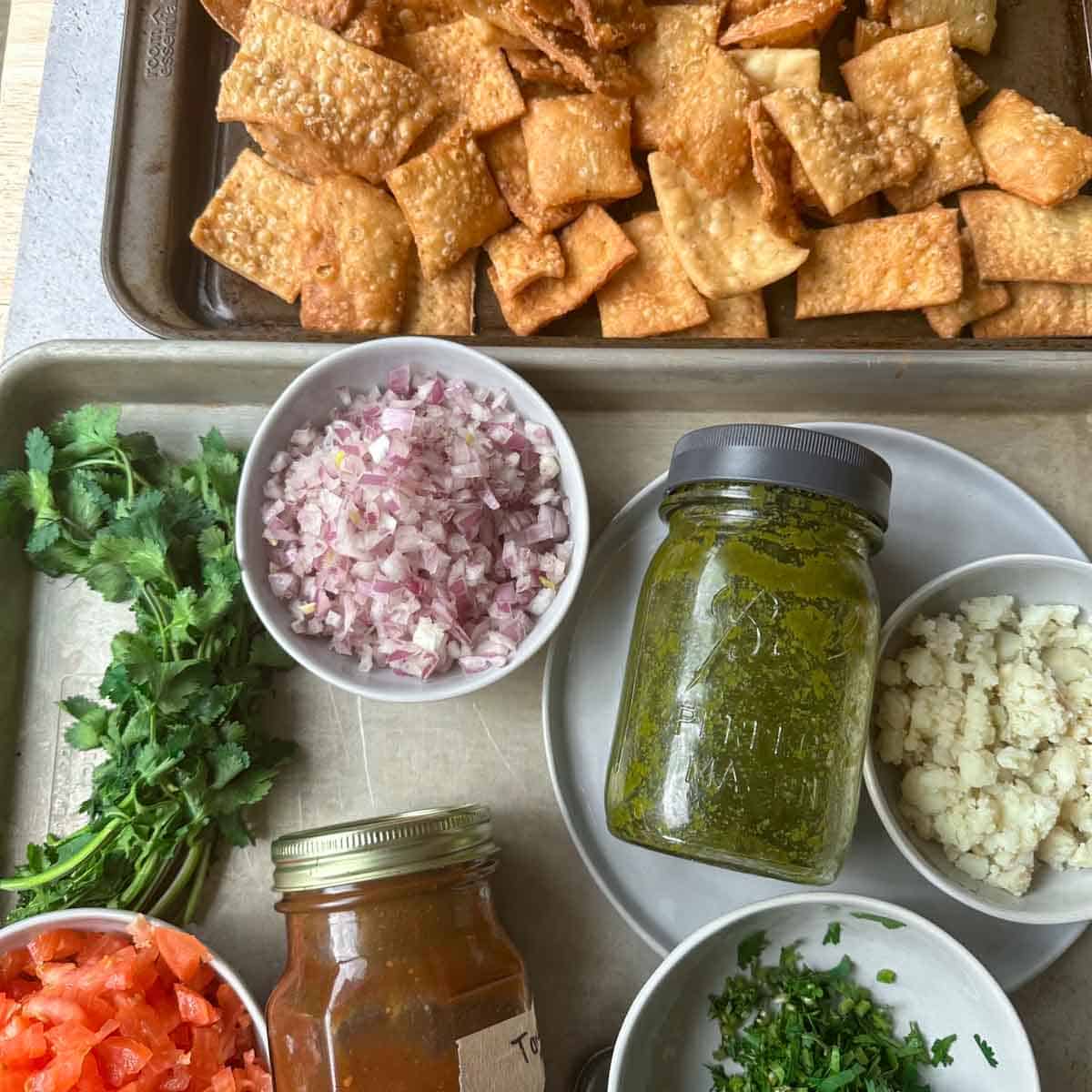 Ingredients of papdi chaat on a sheet tray. From top left to bottom right. Papdi crackers, cilantro, chopped red onion, mint chutney, potatoes, diced tomatoes, tamarind sauce, choped cilantro leaves