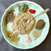 Chaat masala in the middle surrounded by the spices in chaat masala like black salt (kala namak, coriander, cumin, dried mint leaves, chili powder, amchur powder (dried mango powder), asafetida.