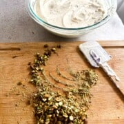 Cutting board with chopped pistachios and bowl in the background with whipped cream and condensed milk