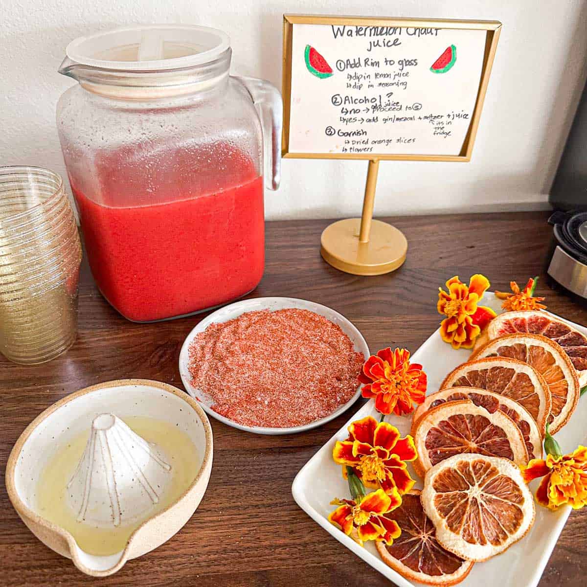 A pitcher of Indian style watermelon juice setup for a party. A sign in the back telling guests how to create a drink along with spices for rim and garnishes.