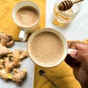 Ginger chai in two cups. Yellow napkins in the background along with honey and fresh ginger root. A brown hand holding one cup of chai in the center.