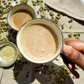 Cardamom Chai. Two cups with cardamom chai. One cup in the center with a brown hand picking it up. Green cardamom pods in the background along with cardamom powder in a small bowl the lower right hand corner of the picture.