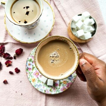Two floral tea cups with rose chai. A brown hand holding one rose chai cup. A small bowl with sugar cubes in the background. Dried rose petals in the lower left of the photo