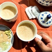 Two cups of decaf masala chai. A brown hand reaching for one cup of chai. Chai masala or spices in the lower left hand corner of the photo. Decaf tea bags in the upper right hand side of the photo along with a blue and white tea pot.