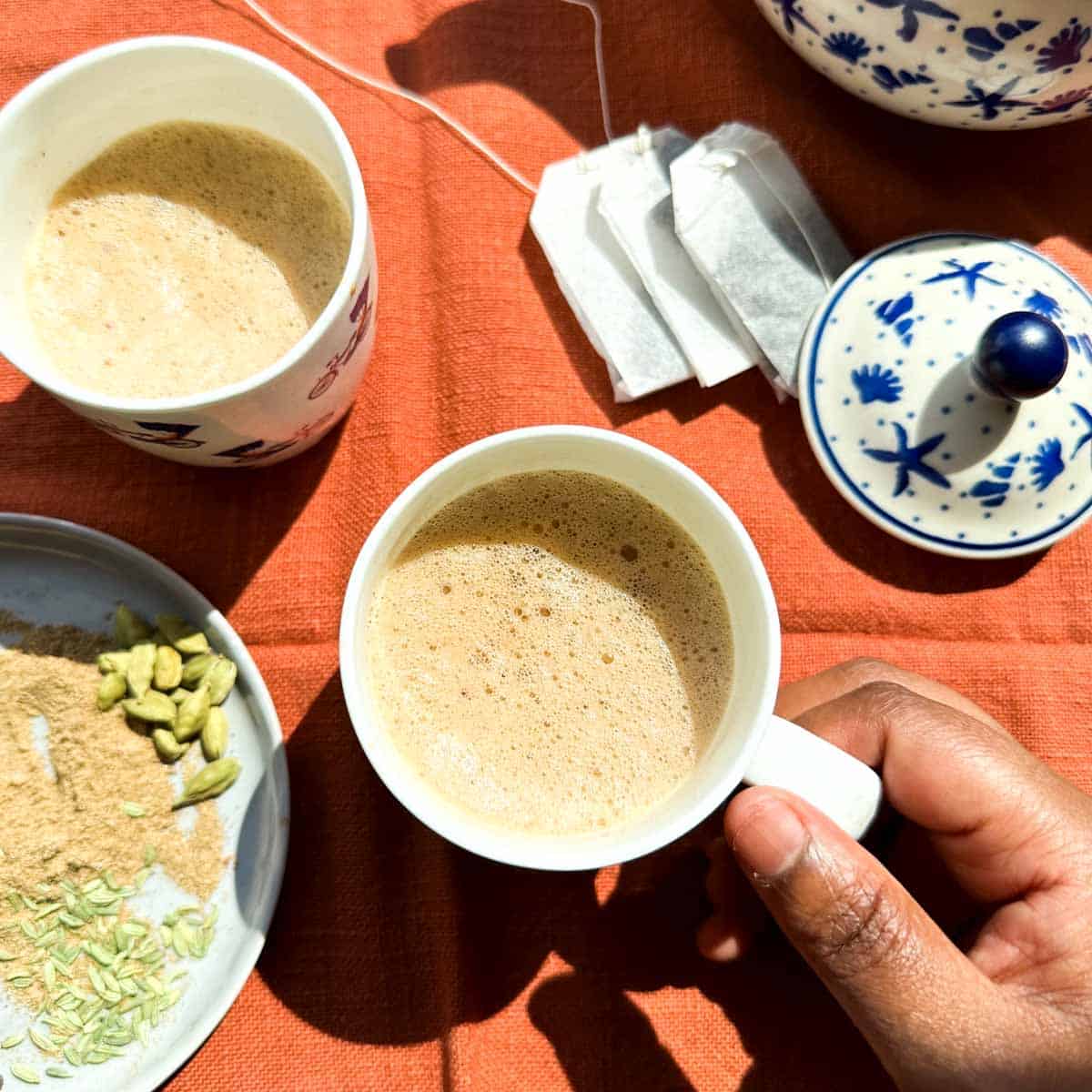 Two cups of decaf masala chai. A brown hand reaching for one cup of chai. Chai masala or spices in the lower left hand corner of the photo. Decaf tea bags in the upper right hand side of the photo along with a blue and white tea pot.