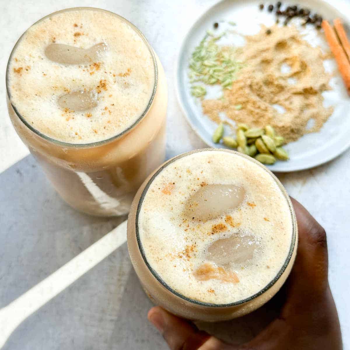 Two tall glasses filled with frothy iced masala chai. A brown hand grabbing one glass of chai. Chai masala or chai spices in the top right hand side of the photo.