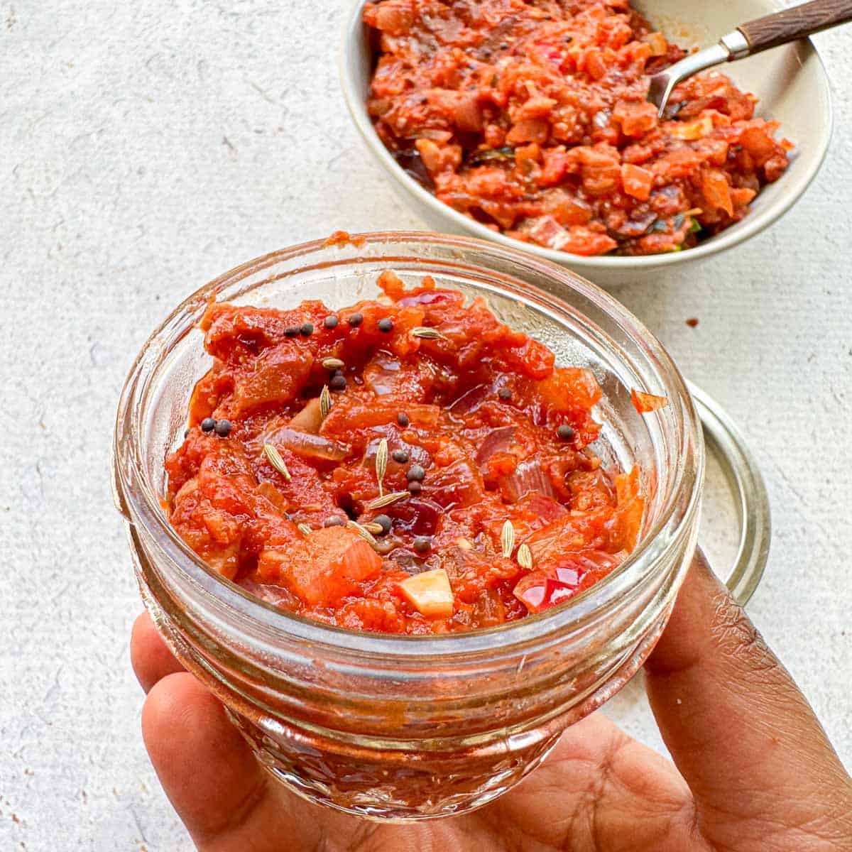 Mason jar with chunky tomato onion chutney it in for storage with a small cup of chutney in the background.