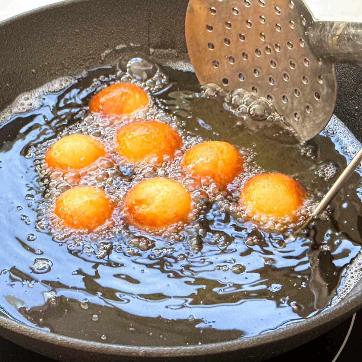 Gulab jamuns being fried with a slotted spoon in oil.
