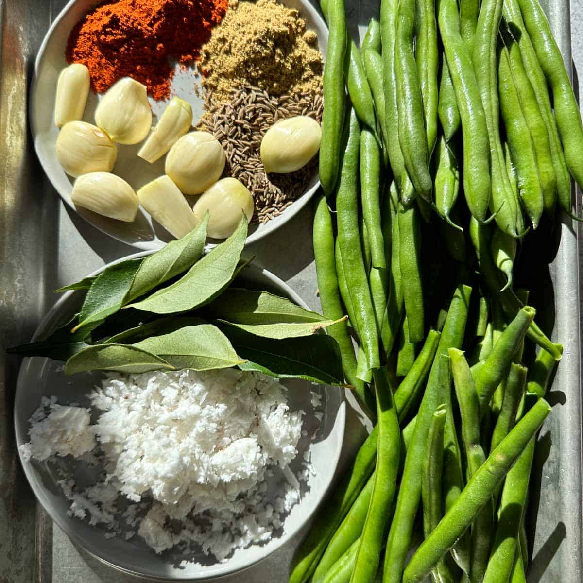 Ingredients on a sheet tray for beans poriyal. Green beans on the right hand side. Garlic, spices, freshly shredded coconut and curry leaves on the left side.