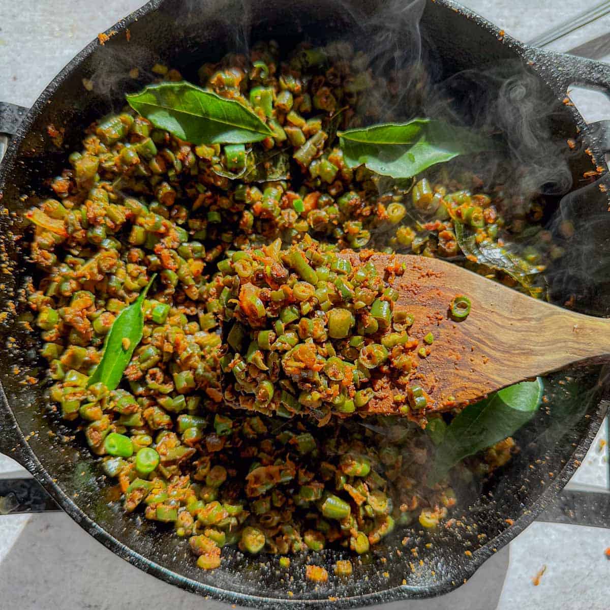 Green beans poriyal in a cast iron pan with a wooden spatula holding some of the beans.