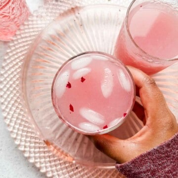 A brown hand holding lychee rose mocktail in a glass with pink hued glasses in the background.