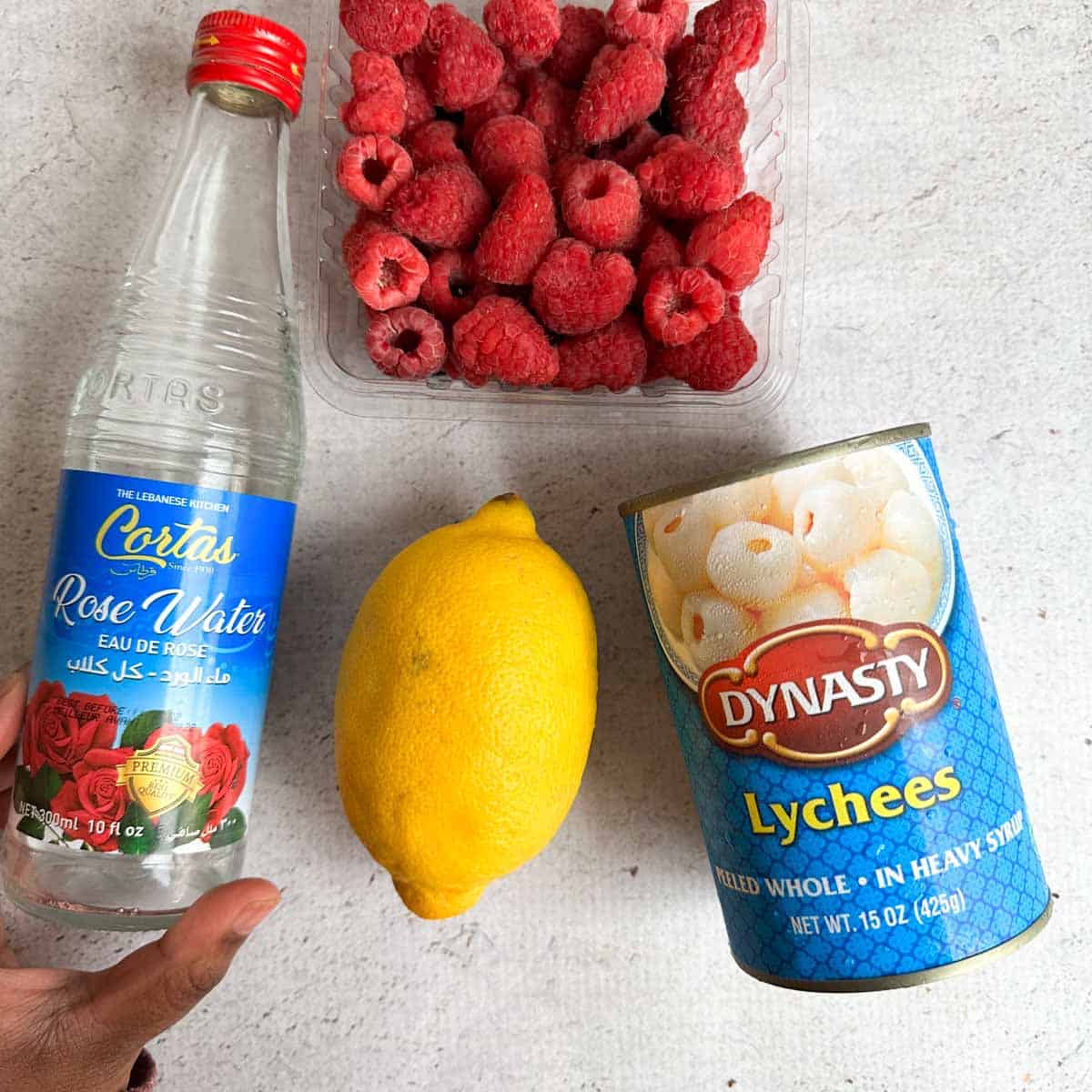 Ingredients for lychee rose mocktail. Rose water in the left, whole lemon in the middle, a can of lychees on the right, and raspberries on top.