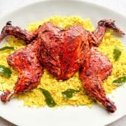 A whole roasted tandoori chicken that is spatchcocked on a large white platter on top of a bed of lemon rice.