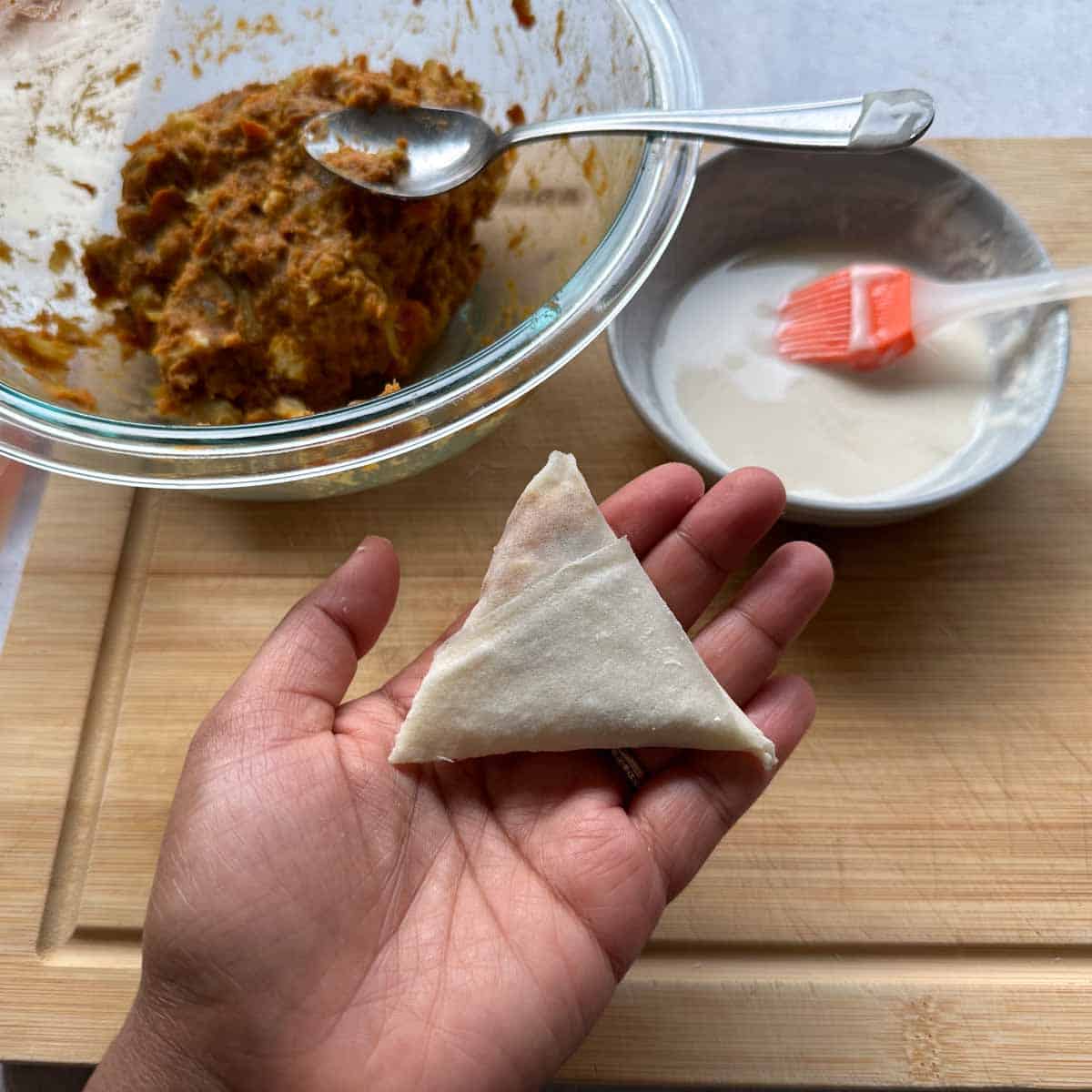 Sweet potato samosa all folded up held in a hand. Flour paste and sweet potato filling in the background.