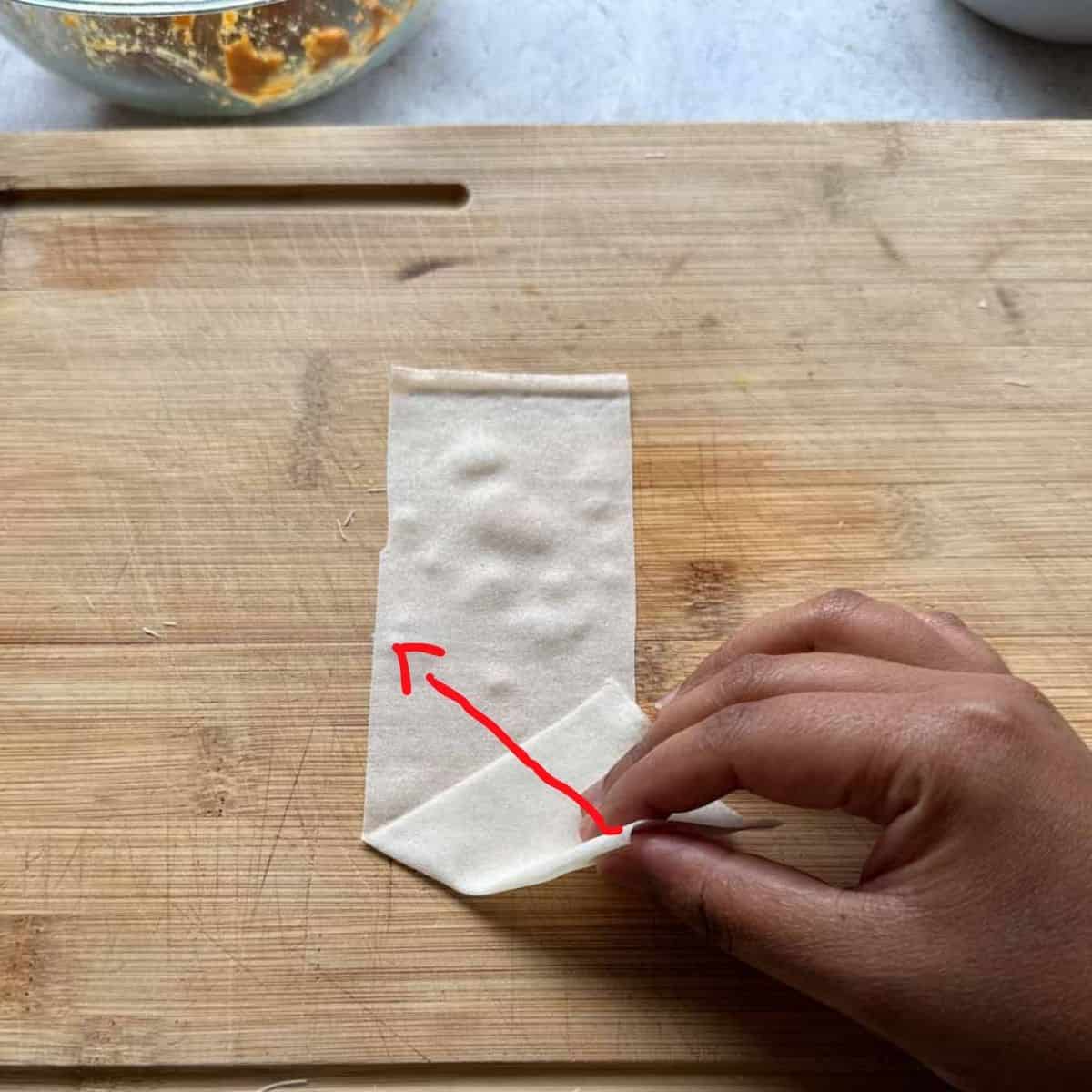 Hand folding a spring roll sheet into the shape of a samosa. A red arrow points to the north west corner of the samosa where the samosa should be folded to.