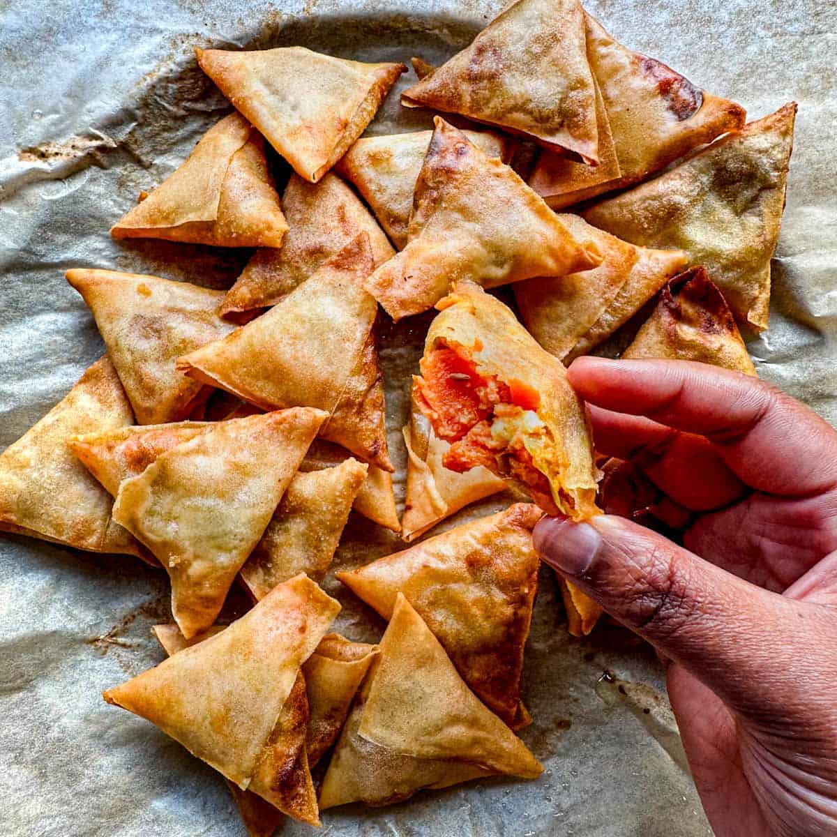 Lots of mini sweet potato samosas on a parchment lined plate. One samosa is torn open with the sweet potato filling showing.