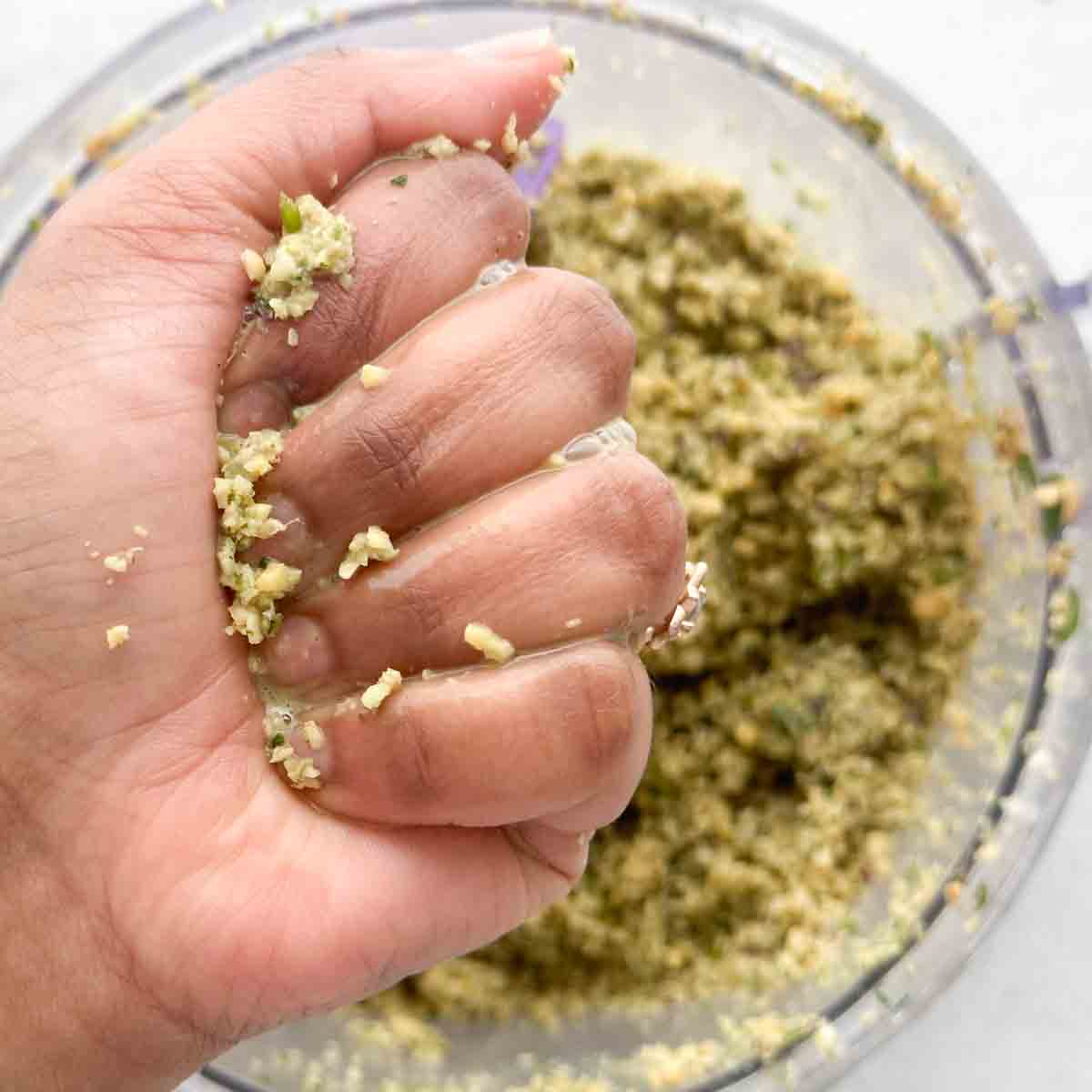 A hand squeezing masala vada mixture with more of the mixture in a food processor in the background.