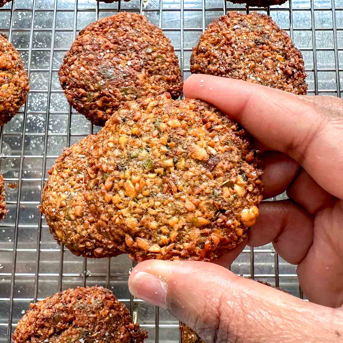 Close up shot with a hand holding a golden masala vada. Other masala vadas on a wire sheet rack.