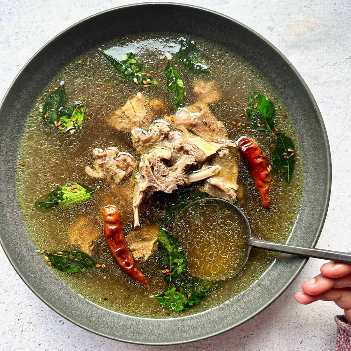 A big gray bowl of mutton bone broth soup with a hand holding a metal spoon.