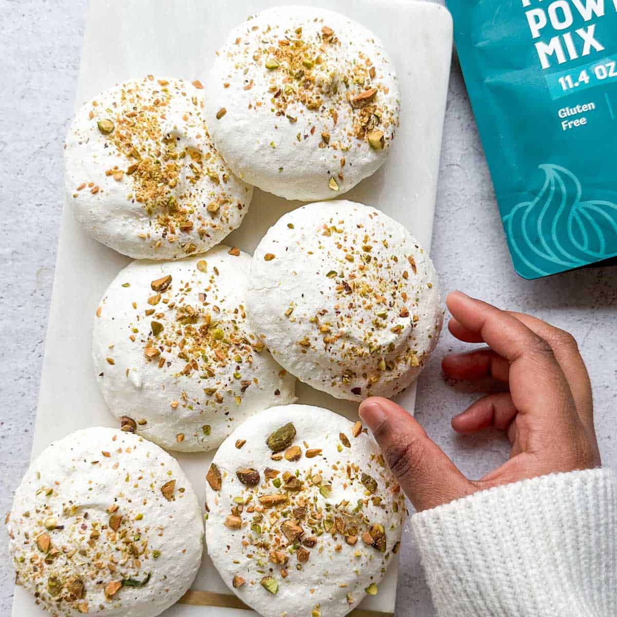 Saffron Pistachio Meringue Cookies on a white tray. A hand is reaching for a cookie and a bag of meringue mix is in the background.