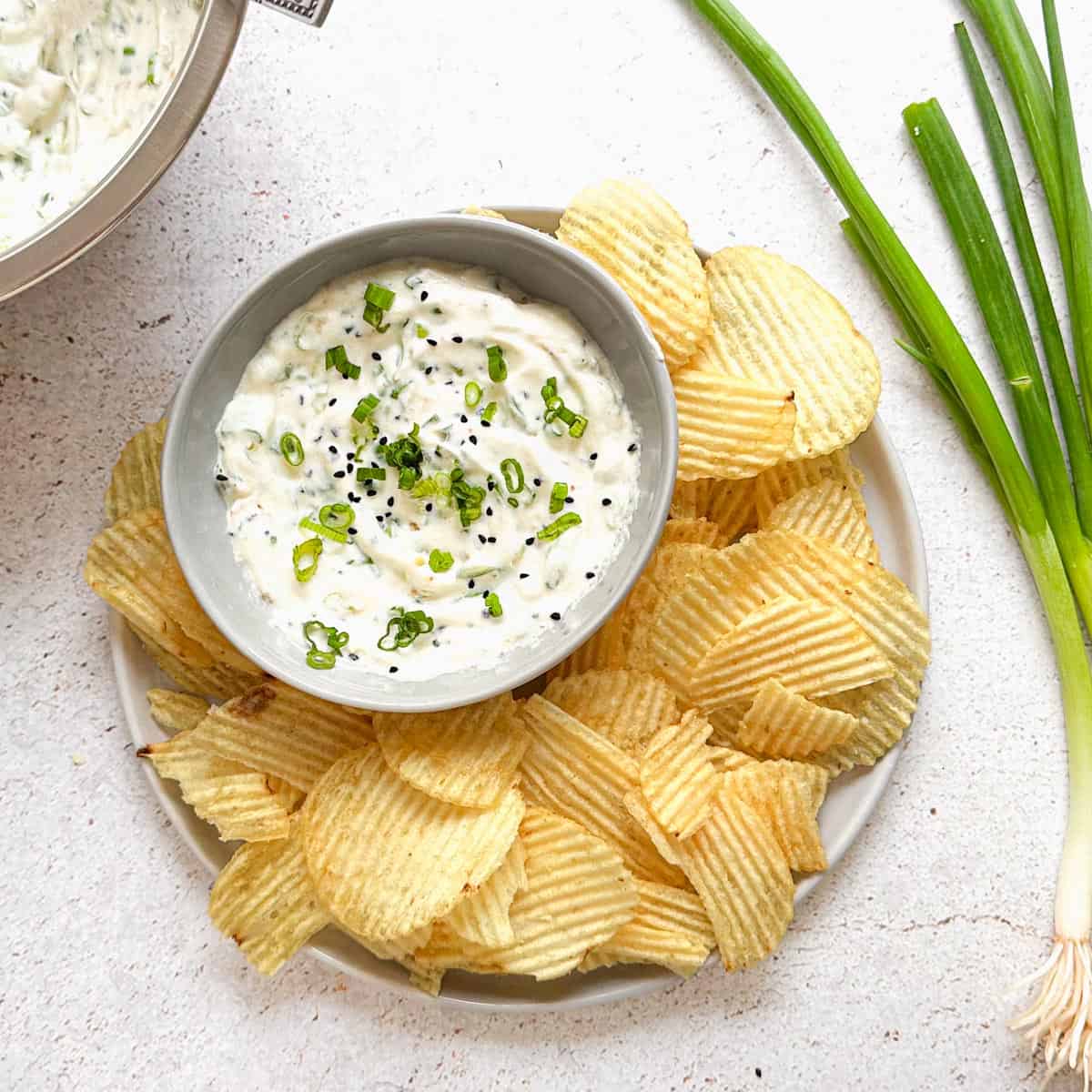 Sour cream and spring onion raita dip in a bowl with wavy potato chips plated along side it. Green onions in the background along with a bigger bowl of dip.