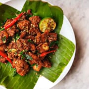 A plate with cooked chili chicken in the center laying on a banana leaf on the exterior.