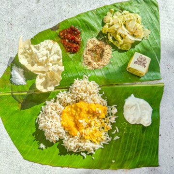 A banana leaf with a South Indian meal on it. The top has salt, applam (papad), indian pickle, milagai podi (gun powder), cabbage poriyal, and a dessert cashew bite. The bottom has rice with dal on top and yogurt on the right side.