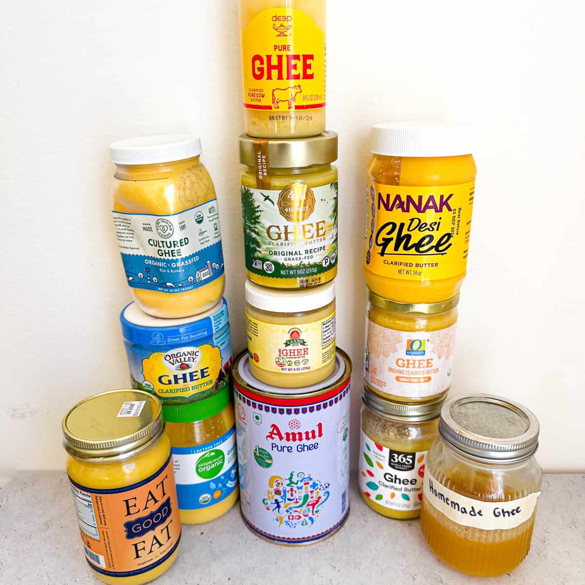 11 different ghee brand jars and 1 homemade jar. The 11 brands from left to right are Ancient Organics, Pure Indian foods, Organic Valley, 4th & Heart, Laxmi Ghee, Amul, Nanak, O Organics, 365 Ghee, and Homemade ghee.