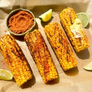 A sheet tray with parchment paper with grilled Indian style corn on the cobs with a spicy masala rub. The tray includes cut limes and a bowl of spicy masala rub. One peice of corn has a big tablespoon of softened butter spread on top.