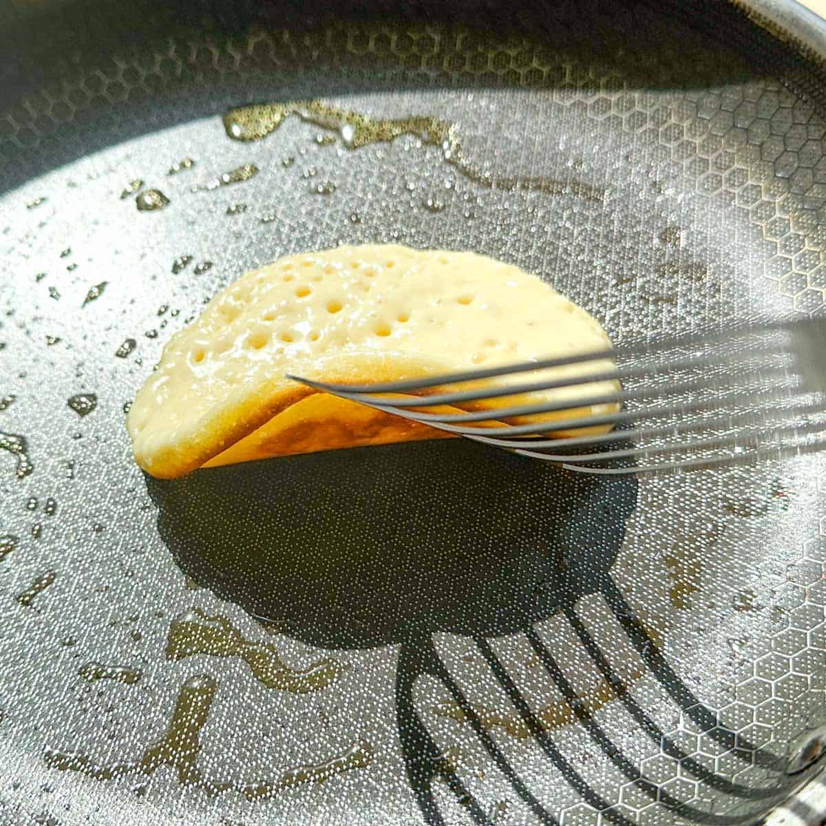 A spatula lifting up a pancake to reveal a golden underside. The top of the pancake has bubbles and is uncooked.