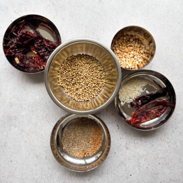Small bowls that contain individual ingredients to make sambar podi or sambar spice blend. One bowl with dried Kashmiri chili pepper, one bowl with chana dal, one bowl with raw white rice and Indian dried red chilis, a big bowl with corainder seeds, and the last bowl has cumin seeds and fenugreek seeds.