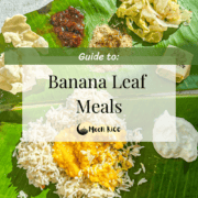 A banana leaf with a South Indian meal on it. The top has salt, applam (papad), indian pickle, milagai podi (gun powder), cabbage poriyal, and a dessert cashew bite. The bottom has rice with dal on top and yogurt on the right side.