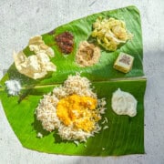A banana leaf with a South Indian meal on it. The top has salt, applam (papad), indian pickle, milagai podi (gun powder), cabbage poriyal, and a dessert cashew bite. The bottom has rice with dal on top and yogurt on the right side.
