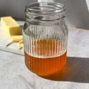 Ghee in a glass mason jar in the forefront. Stick of butter on a white board in the background.