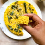 A brown hand holding a cross section of nylon khaman dhokla. The cross section shows big air bubbles and a spongy dhokla. The background shows the rest of the dhokla on a serving plate along with cilantro-mint green chutney and chai that are served with the dhokla.