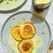 Rasmalai pancakes on two plates with saffron milk in a pitcher.