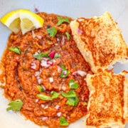 Pav bhaji plated in a bowl. The pav or dinner roll bread is toasted and on the side. The bhaji or vegetable and potato curry has raw onions, cilantro, and lemon served on top as garnish.