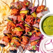 Charred chicken tikka kebabs on wooden skewers. The kebabs are on a white plate accompanied with naan, green chutney, and raw red onions. The skewers have red onions, chicken, and then green bell peppers on them.