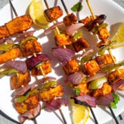 Tandoori paneer tikka skewers on a plate with lemon and cilantro as garnish. The skewers are wooden skewers with red onion, marinated paneer, green bell pepper alternating on the kebab. The veggies and paneer have a slight char on them.