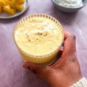 High protein mango lassi in a glass with a hand holding it. Two bowls behind the mango lassi that have mango chunks and greek yogurt.