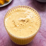 High protein mango lassi in a glass with a hand holding it. A plate behind the mango lassi that has mango chunks and a bowl that has greek yogurt.