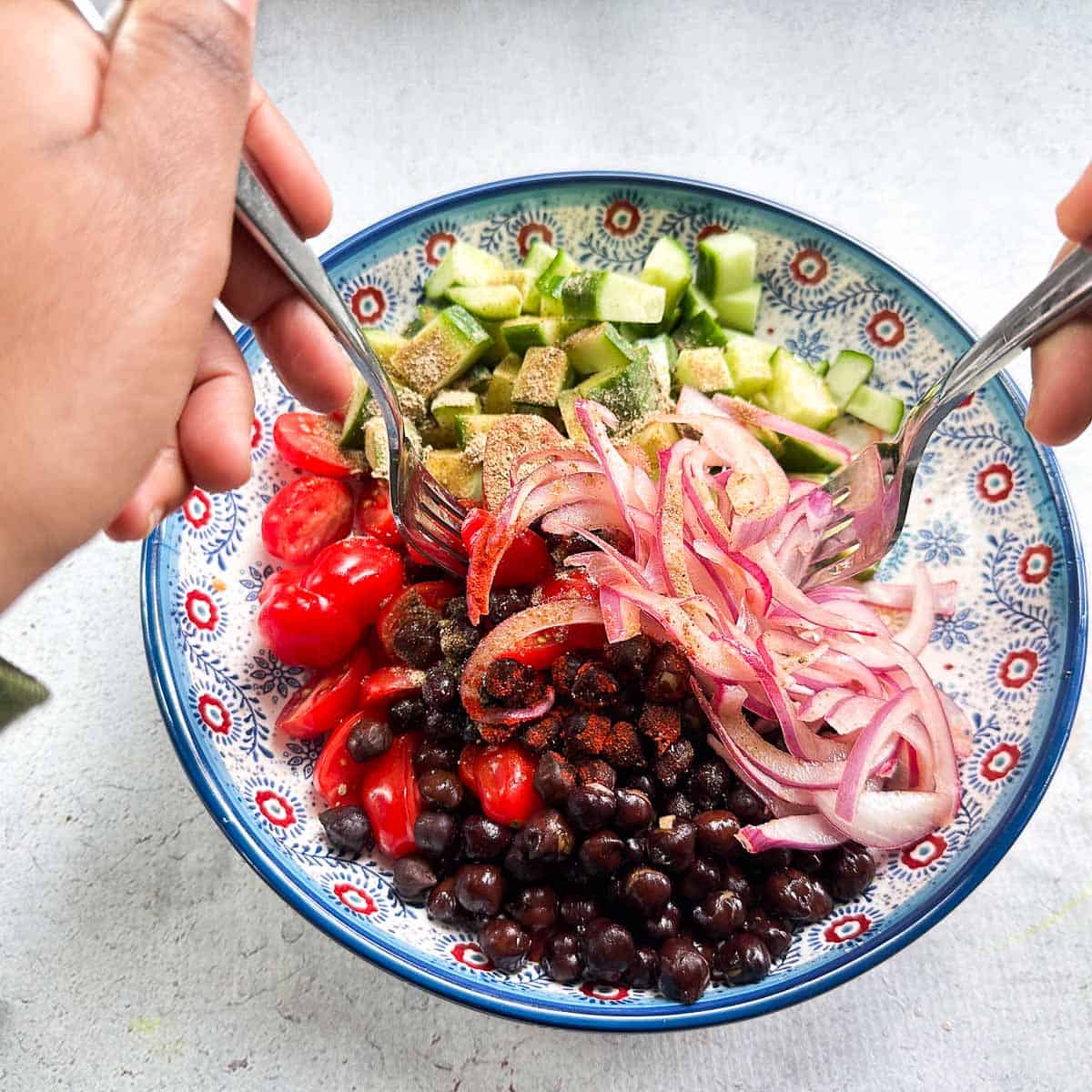 A bowl filled with a kala chana or chickpea salad that is being tossed. The salad has black chickpeas, pickled onions, cucumbers, tomatoes and spices that are being tossed with two forks.