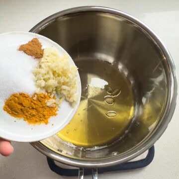 Turmeric, cinnamon, finely chopped garlic, and salt being added to a pot with melted ghee and whole green cardamom pods.