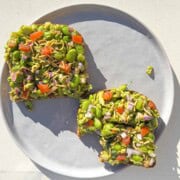 Two slices of edamame chaat avocado toast. The toast has shelled edamame, cut red onions, tomatoes, green bell pepper, cilantro, salt, chaat masala, and thin sev on top.