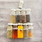 10 essential Indian spices laid out in their spice containers. The spices from bottom left to the top are green cardamom, Kashmiri chili powder, turmeric powder, cumin seeds, chaat masala, coriander seeds, black peppercorn, cinnamon sticks, mustard seeds, and asafetida (hing).